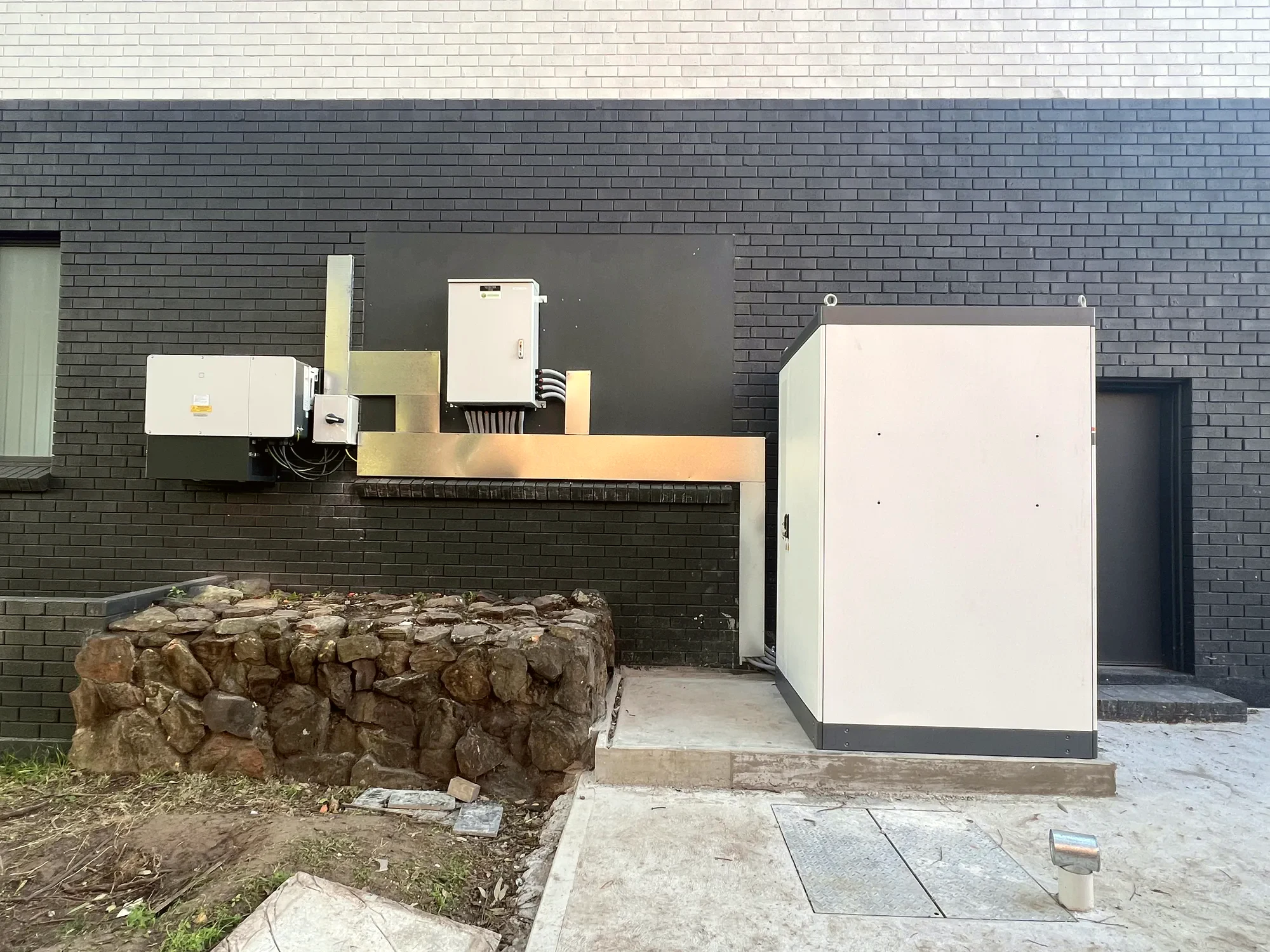 Battery enclosures and an inverter mounted on an exterior brick wall, with the ground works finished around the pad.