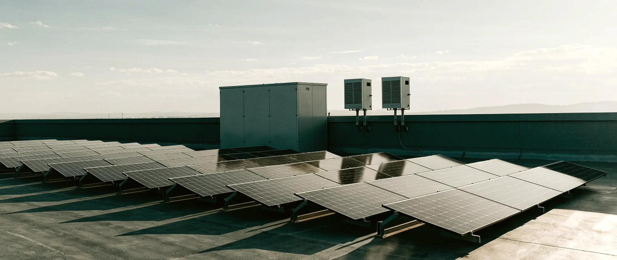 A rooftop solar array in the foreground with a battery cabinet and two inverters standing at the parapet edge behind it.