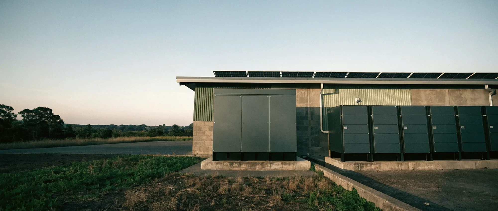 A battery energy storage cabinet and inverter bank on a concrete pad outside a light industrial building, with a rooftop array along the roofline behind.