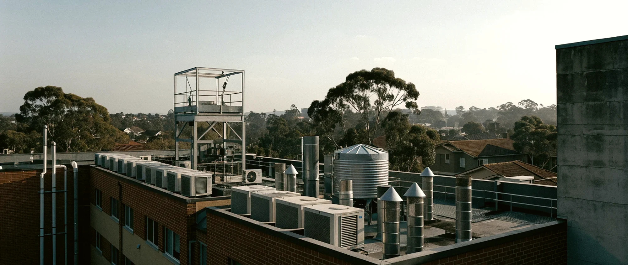 The roof of a brick apartment building, crowded with air conditioning plant, vent stacks, a water tank and a lift overrun, with suburban rooftops and gums beyond.