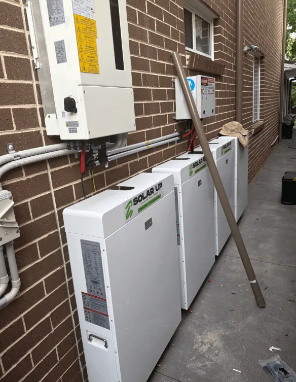 Four Solar Up branded wall batteries in a row on a red brick wall, with the inverter and isolators mounted above them.