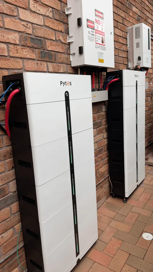A white Pytes battery stack on red brick paving beneath a Dyness enclosure and an isolator, with the DC cabling run neatly to the wall.