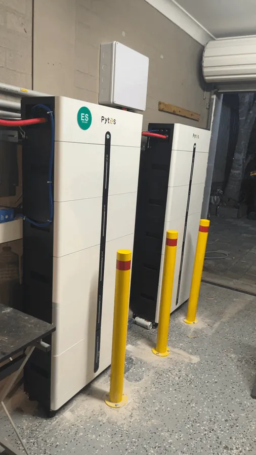 A white and a black Pytes battery stack standing side by side in a carport, protected by red and yellow impact bollards.