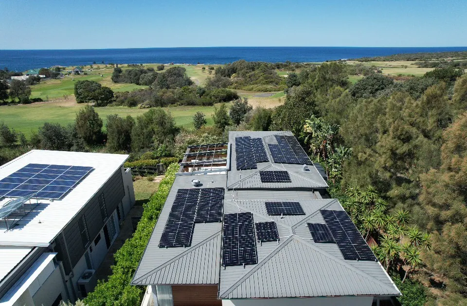 A drone view of solar arrays across two adjoining roofs, with parkland and the ocean on the horizon.
