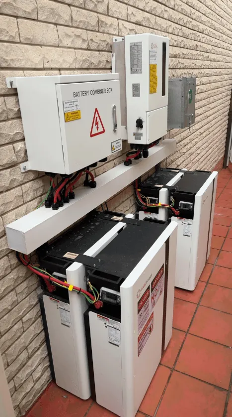 A battery combiner box and an inverter mounted on a cream brick wall, with four battery modules on the terracotta tiles below and the DC cabling landed into the box above.