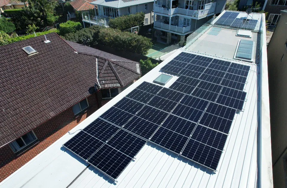 A drone view of a solar array laid out along the flat roof of a house, with a tiled roof and gardens beside it.