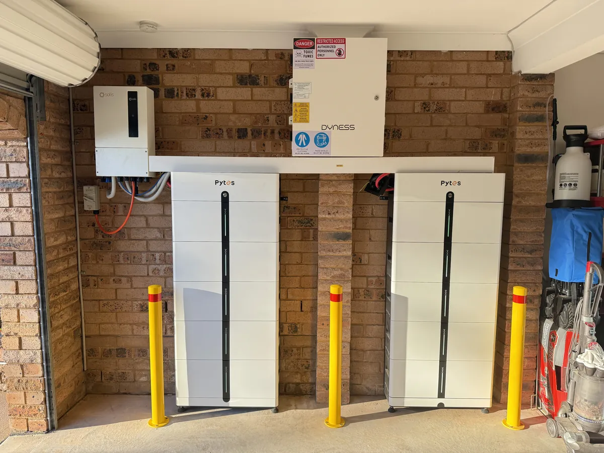 A garage wall with a Solis inverter and a Dyness combiner box mounted on brick, and two white Pytes battery stacks standing below behind yellow impact bollards.