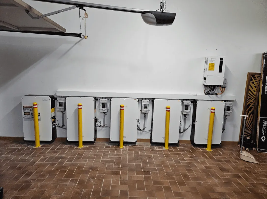 Six wall-mounted batteries and their isolators running the length of a garage wall above a herringbone brick floor, with the inverter at the far end.