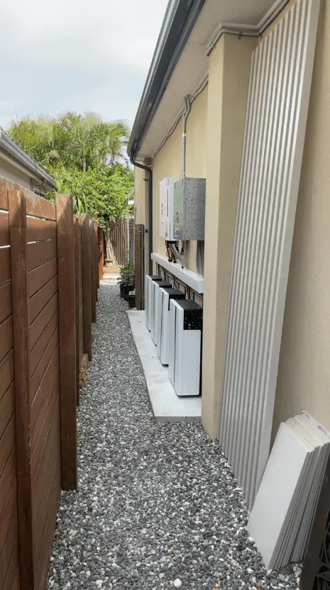 A run of wall batteries and their isolators down the narrow gravel side passage of a house, seen from the far end.