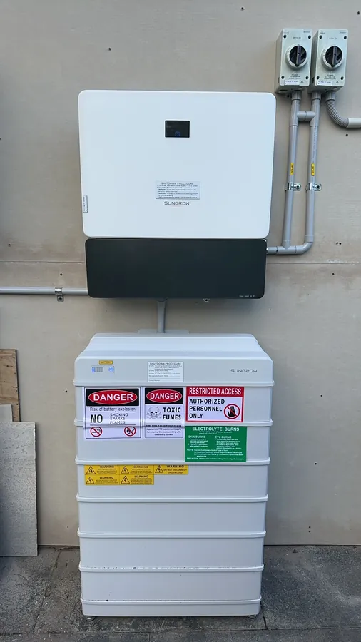 An inverter mounted above a wall battery on a rendered exterior, with the switchboard isolators alongside and the required safety signage across the battery.