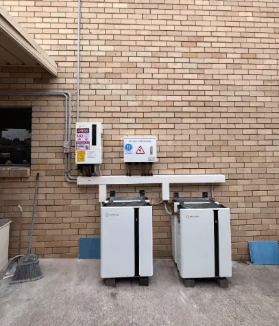 Two white wall batteries standing side by side on a brick wall beneath a mounting rail, with the cabling dropped into each unit.