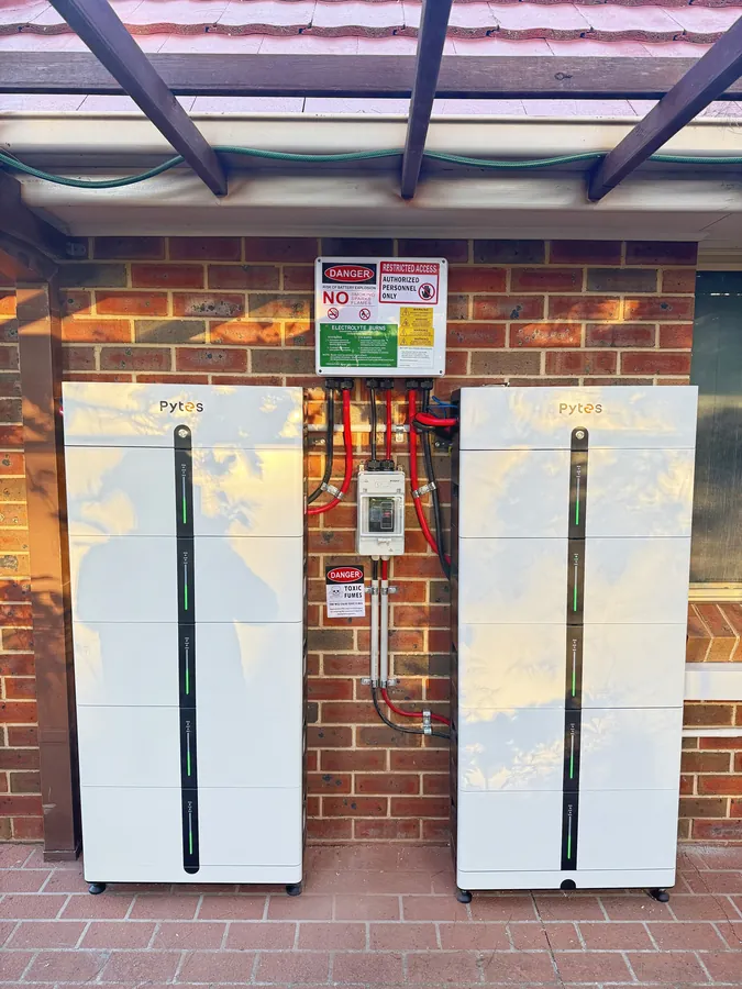 Two white Pytes battery stacks either side of an isolator on a red brick wall, under a timber pergola, with the sky reflected in their front panels.