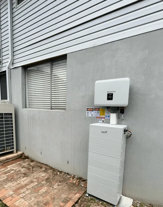 An inverter and a battery mounted on the rendered side wall of a house, next to the air conditioning condenser, above brick paving.