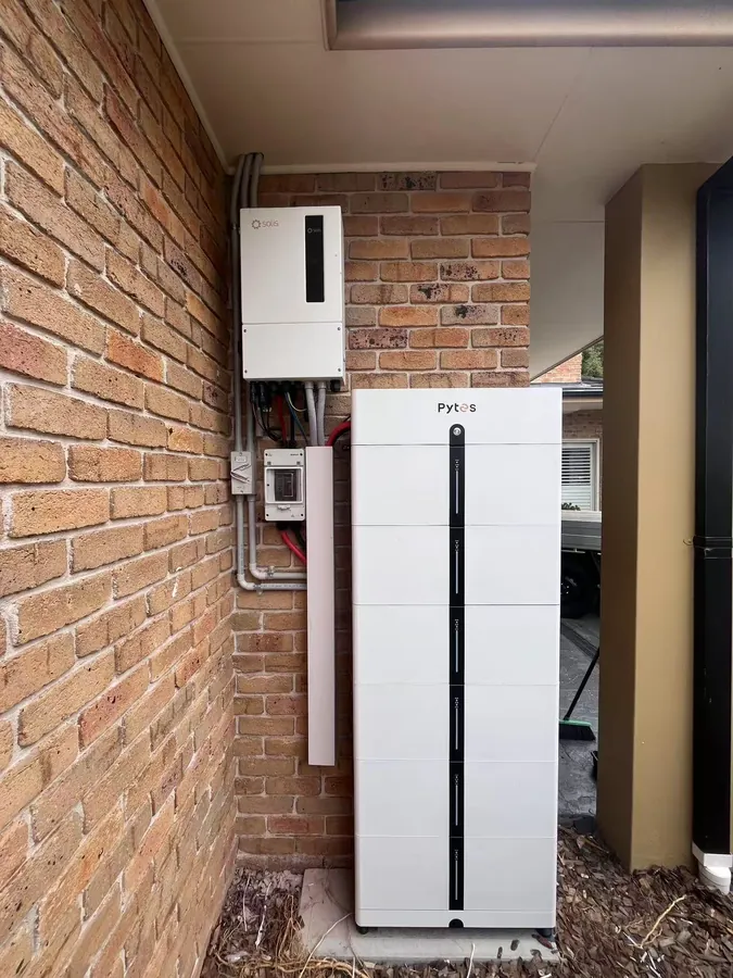 A single white Pytes battery stack and a Solis inverter mounted on face brick in the narrow side passage of a house.