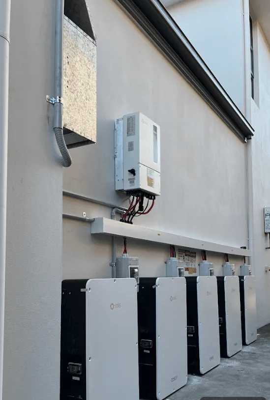 A run of wall-mounted batteries and their isolators along the rendered side of a building, with the inverter and switchboard at the near end.