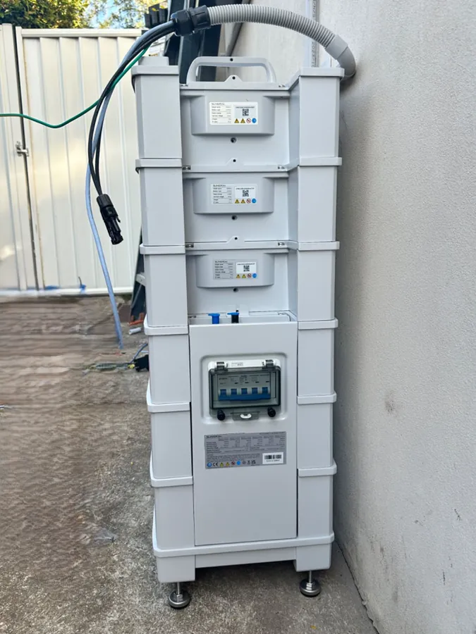A three-module battery stack against a rendered wall, with the DC isolator and its enclosure landed at the base and the conduit run to the wall.