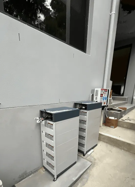 Two battery stacks standing against a rendered wall beside a concrete step, with the isolators and cabling run above them.