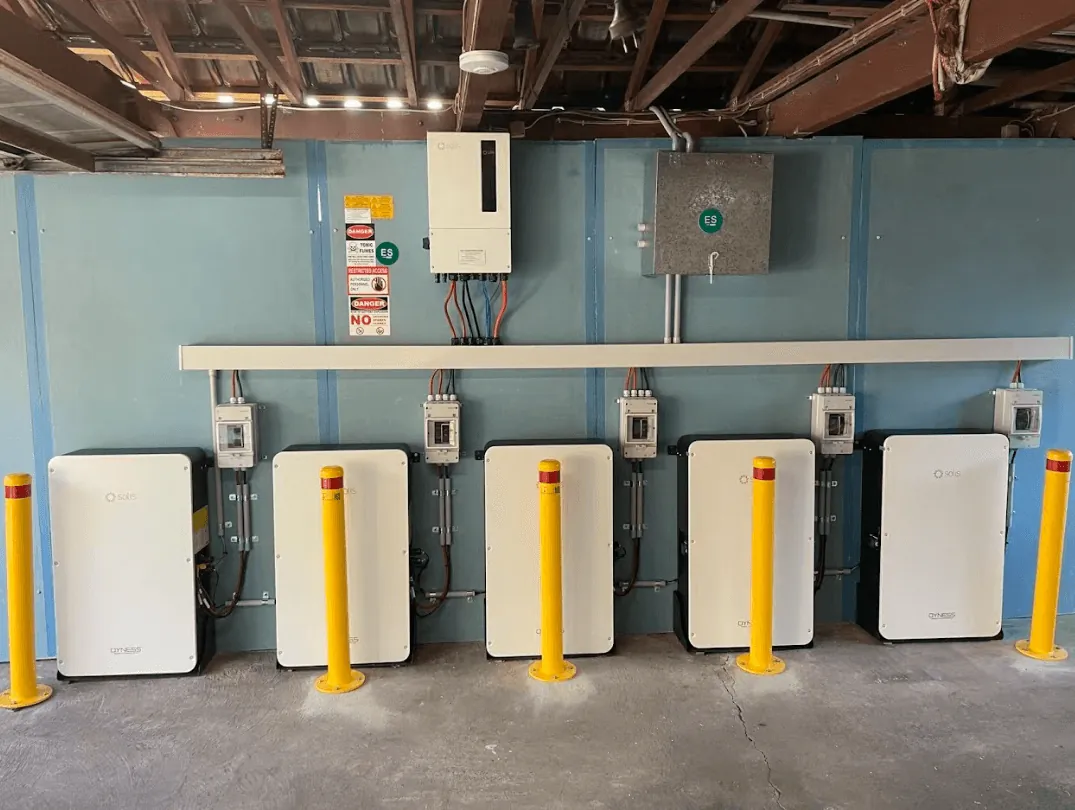 Five white wall-mounted batteries in a line under a house, each with its own isolator above, an inverter and metering box on the wall, and yellow bollards along the front.
