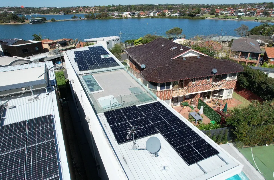A drone view of a solar array along a flat roof, with a bay and further housing behind it.