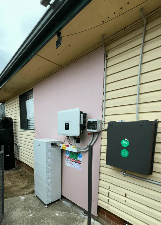 An inverter, a battery and a black enclosure mounted on the weatherboard side wall of a house.